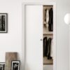 The ECLISSE Classic Single Pocket Door Kit is partially open, revealing hanging coats and jackets inside. The space features minimal decor, light walls, framed art resting on the floor, and a striped bedspread for a clean, modern look.