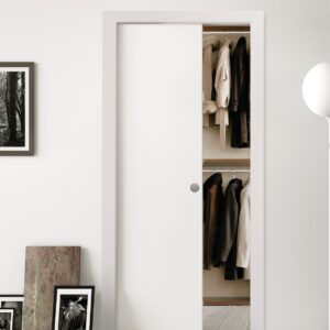 The ECLISSE Classic Single Pocket Door Kit is partially open, revealing hanging coats and jackets inside. The space features minimal decor, light walls, framed art resting on the floor, and a striped bedspread for a clean, modern look.