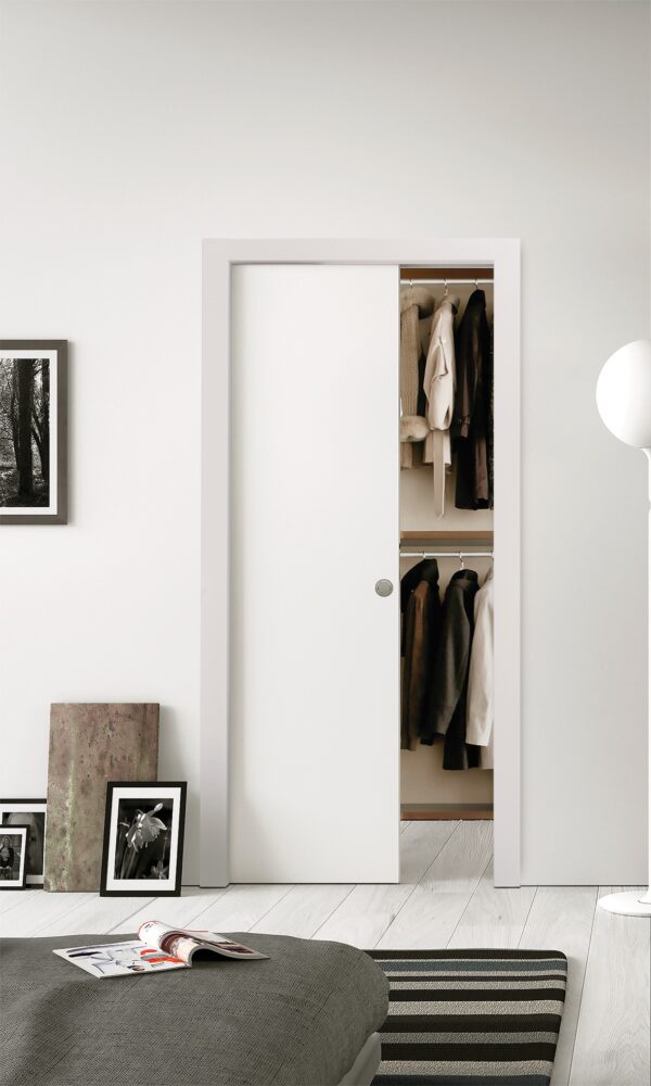 The ECLISSE Classic Single Pocket Door Kit is partially open, revealing hanging coats and jackets inside. The space features minimal decor, light walls, framed art resting on the floor, and a striped bedspread for a clean, modern look.