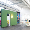 A spacious art studio with high windows features a green wall fitted with the ECLISSE Pocket Door System – Syntesis® Single Wiring-Ready, glowing lights, colourful bags in the corner, and scattered paintings, art supplies, and stools throughout the bright room.