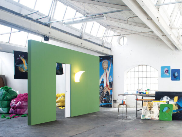 A spacious art studio with high windows features a green wall fitted with the ECLISSE Pocket Door System – Syntesis® Single Wiring-Ready, glowing lights, colourful bags in the corner, and scattered paintings, art supplies, and stools throughout the bright room.