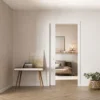 Minimalist interior with light beige walls, wooden floor, a small mid-century desk with art prints, a potted plant in a wicker basket, and a view into a cosy bedroom through an ECLISSE Classic Single Pocket Door Kit.