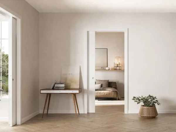 Minimalist interior with light beige walls, wooden floor, a small mid-century desk with art prints, a potted plant in a wicker basket, and a view into a cosy bedroom through an ECLISSE Classic Single Pocket Door Kit.