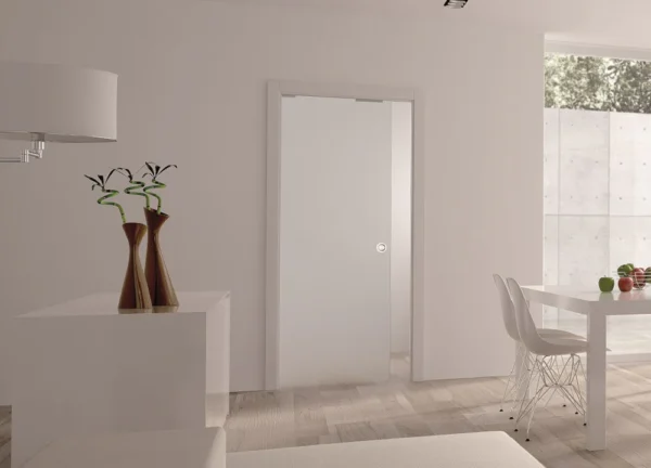A modern, minimal room with light walls and floors features an ECLISSE Pocket Door System - Classic Single Plain Satin 8mm Glass with Stainless Steel Round Handle, a white dining table with clear chairs, fruit on the table, and two vases with abstract branches.