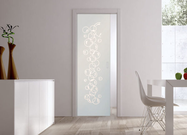 An ECLISSE Pocket Door Systems Classic Single Glass Door with frosted circles enhances a minimalist white room featuring a cupboard, dining table, white chair, decorative vases, and natural light from a window.