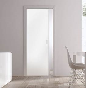 An Eclisse Pocket Door System - CLASSIC SINGLE PLAIN GLASS SATIN 8mm 826X2040mm is partially open in a minimalist room with light walls, wooden flooring, a white chair, a white cupboard, and natural light streaming from the right window.
