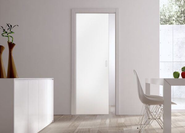 A modern, minimalist room with an ECLISSE Pocket Door System – Classic Single Glass Plain Satin 8mm, a sleek white cupboard topped with two tall vases, and a dining table with a wire chair. Natural light fills the space through a large window.