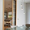 A modern dining room with a glass table and pendant lights leads to a cosy kitchen with a round table, separated by an ECLISSE Pocket Door System - Classic Single Wiring-Ready featuring frosted glass panels.