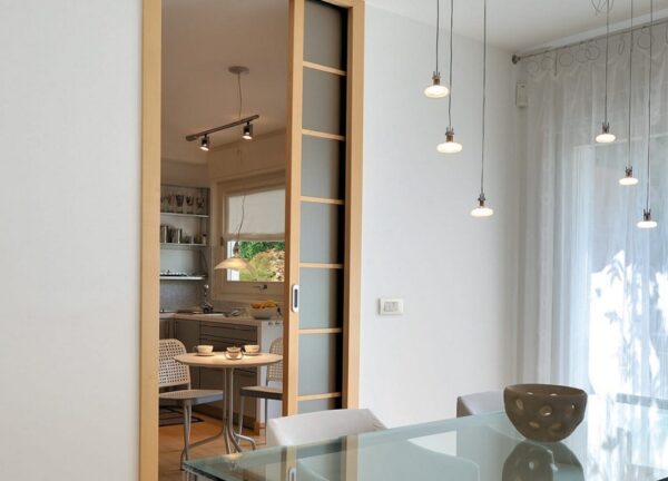 A modern dining room with a glass table and pendant lights leads to a cosy kitchen with a round table, separated by an ECLISSE Pocket Door System - Classic Single Wiring-Ready featuring frosted glass panels.