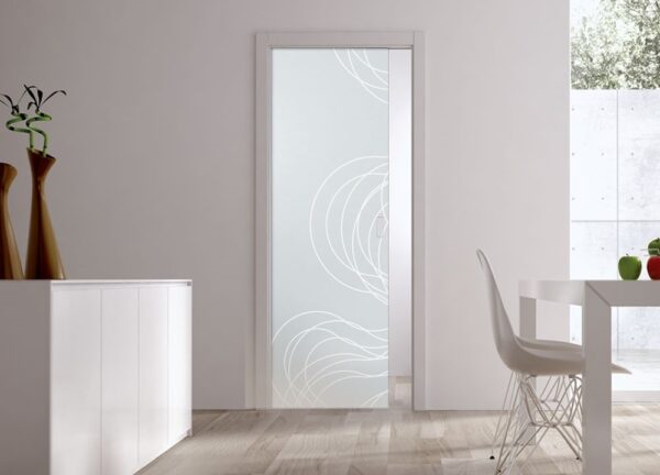 A modern room with light wood flooring, a white sideboard, two tall vases, and an ECLISSE Pocket Door Systems - Classic Single Glass Door with a white wavy pattern. A white dining table and chair sit in natural light from outside.
