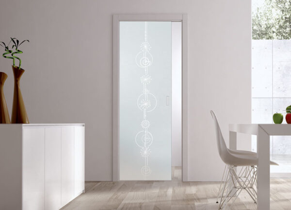 A modern interior showcases ECLISSE Pocket Door Systems—Classic Single Glass Doors 10mm (plain or patterned), frosted with geometric designs, light wood floors, a white cupboard with tall vases, and a white dining table with a wire chair by a large window.