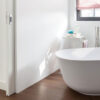 A modern bathroom features a white freestanding bath, wooden flooring, and a red and white striped window blind. An adjacent bedroom is seen through an ECLISSE Pocket Door System - Classic Double Wiring-Ready, adding style and saving space.