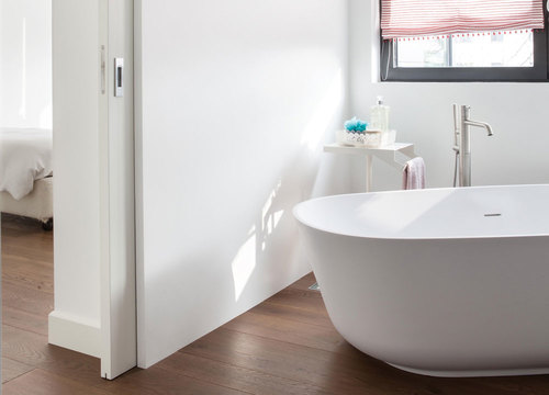 A modern bathroom features a white freestanding bath, wooden flooring, and a red and white striped window blind. An adjacent bedroom is seen through an ECLISSE Pocket Door System - Classic Double Wiring-Ready, adding style and saving space.