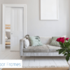 A bright, modern living room with white floors, a light grey sofa, and red roses in a vase. An open Rocket Door Frames - 70mm Pocket Door Kit shows a glimpse of a bed. The Rocket Door Frames logo appears in the bottom left corner.