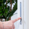 A hand with a silver bracelet presses a small lever on the AURA Round Flush Pull Handle Set for sliding pocket doors, fitted on a white door, with green leafy plants blurred in the background.