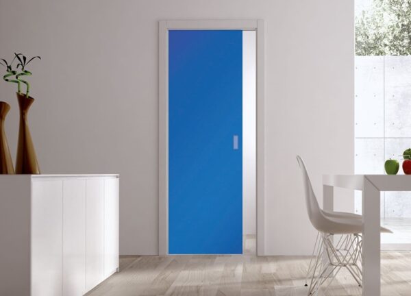 A modern room with light wood floors, white walls, and an ECLISSE Pocket Door Systems - Classic Single Coloured Glass Door 10mm in blue. White cupboards on the left display two tall vases; a white dining table and chair are on the right.