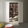 A modern room with curved walls features sleek Eclisse Pocket Door System - CURVED DOUBLE doors that open to a cosy reading nook with bookshelves, armchair, and ottoman. Colourful poufs are in the foreground; the dining area is visible in back.