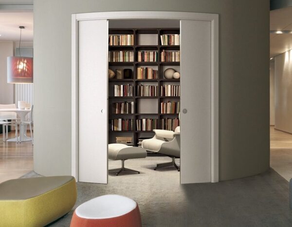 A modern room with curved walls features sleek Eclisse Pocket Door System - CURVED DOUBLE doors that open to a cosy reading nook with bookshelves, armchair, and ottoman. Colourful poufs are in the foreground; the dining area is visible in back.
