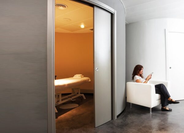 A woman sits on a white chair in a modern waiting area, viewing a tablet. Beside her, an ECLISSE Pocket Door System - Curved Single is open, revealing a massage table in a softly lit, clean, minimalist treatment room.