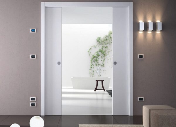 An ECLISSE Pocket Door System - Classic Double Wiring-Ready leads to a minimalist bathroom with a white bath, wooden stool, and green vine on the wall. Modern lighting and control panels complete the sleek, refined look.