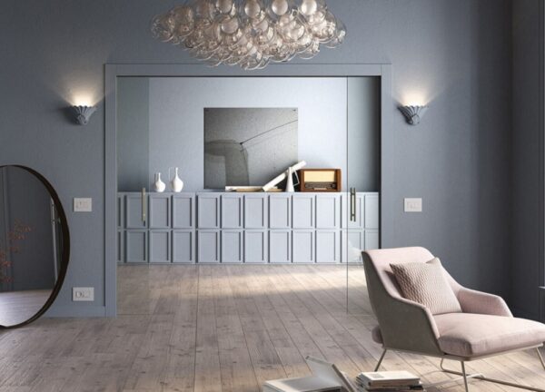 A modern living room with grey walls and light wood floors features a pink armchair, a large round mirror, a glass bubble chandelier, and the ECLISSE Pocket Door System - Classic Double Wiring-Ready as stylish sliding doors.