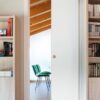 Bookshelves filled with books flank a sleek ECLISSE Classic Single Pocket Door Kit, partially open to reveal two green chairs and a desk in a bright, modern room with exposed wooden ceiling beams.