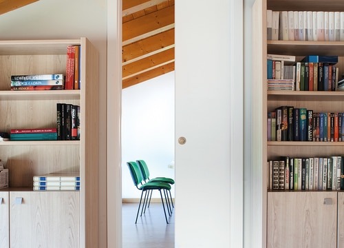 Bookshelves filled with books flank a sleek ECLISSE Classic Single Pocket Door Kit, partially open to reveal two green chairs and a desk in a bright, modern room with exposed wooden ceiling beams.