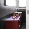 A modern bathroom features dark tiled walls, a red vanity unit with a white basin, a small basket, and a reed diffuser. Light enters from two windows above. Space is maximised by the entrance with the ECLISSE Pocket Door System - Syntesis® Single.