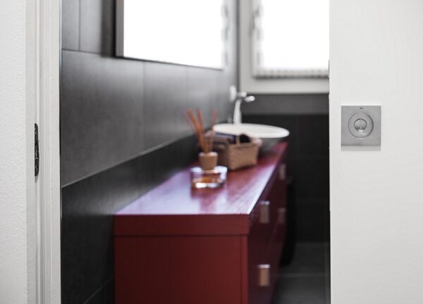 A modern bathroom features dark tiled walls, a red vanity unit with a white basin, a small basket, and a reed diffuser. Light enters from two windows above. Space is maximised by the entrance with the ECLISSE Pocket Door System - Syntesis® Single.