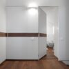 A minimalist hallway with white walls, wood flooring, and a dark wood accent features an ECLISSE Syntesis® Hinged Single With Door. The open door reveals a bathroom with white fittings; a wall-mounted light is above the doorway.