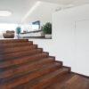 A modern interior with wide wooden stairs opens to a bright living area. White walls, green plants, and the ECLISSE Door system Syntesis® Hinged Single With Door create a contemporary, clean, and open atmosphere.