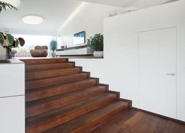 A modern interior with wide wooden stairs opens to a bright living area. White walls, green plants, and the ECLISSE Door system Syntesis® Hinged Single With Door create a contemporary, clean, and open atmosphere.