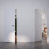 Two white ECLISSE Door system - Syntesis® Hinged Single With Door units in a minimalist room: one is slightly open to greenery, the other fully open reveals a cosy space with fireplace, mirror, modern light fitting, and chair.