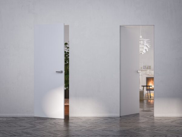 Two white ECLISSE Door system - Syntesis® Hinged Single With Door units in a minimalist room: one is slightly open to greenery, the other fully open reveals a cosy space with fireplace, mirror, modern light fitting, and chair.
