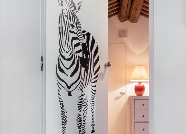 A white ECLISSE Door system - Syntesis® Hinged Single With Door, featuring a black and white zebra illustration, is partially open to a small room with a red lamp on a white chest of drawers and exposed wooden ceiling beams.