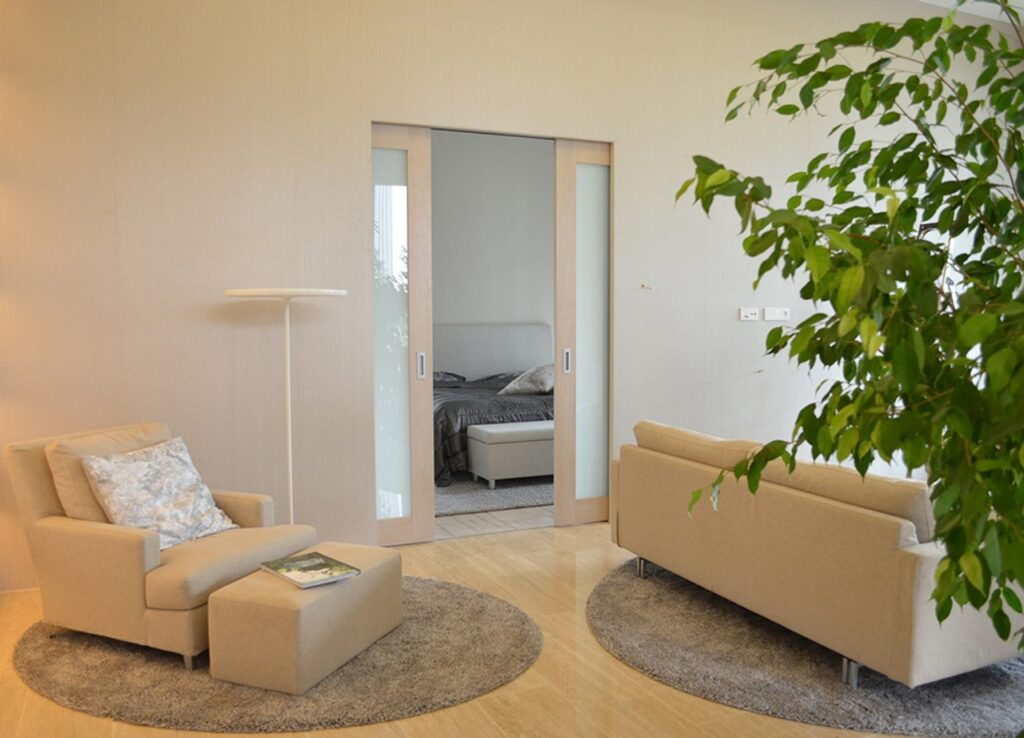 A modern living room with beige armchairs, a sofa, circular rugs, a leafy plant, and a standard lamp. ECLISSE Pocket Door System - Syntesis Double Glass reveals a glimpse of a neatly made bed in the adjoining bedroom.
