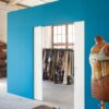 A tailor’s studio with a bright blue ECLISSE Pocket Door System - Syntesis® Double, shelves and colourful fabric rack, plus a vintage dressmaker’s dummy in a decorative corset. Sunlight streams in through the window.