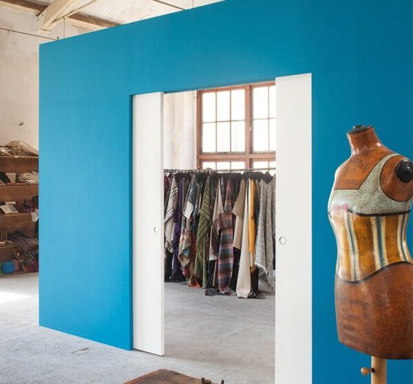 A tailor’s studio with a bright blue ECLISSE Pocket Door System - Syntesis® Double, shelves and colourful fabric rack, plus a vintage dressmaker’s dummy in a decorative corset. Sunlight streams in through the window.