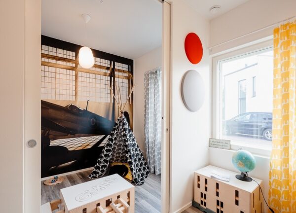 A bright children’s room features an ECLISSE Pocket Door System - Syntesis® Double for easy access, a teepee tent, table and stools, globe, colourful wall decor, a large window with yellow curtains, and an aeroplane mural on the back wall.