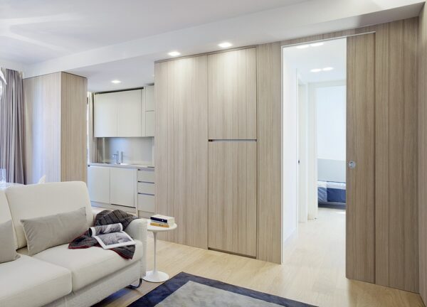 A modern flat features light wood panelling, an open kitchen with white cupboards, and a beige sofa. Hallway-to-bedroom access uses the ECLISSE Pocket Door System - Syntesis® Single for a seamless, minimalist look.