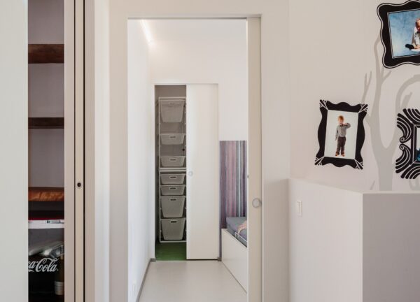 A bright, modern hallway with white walls features the ECLISSE Pocket Door System - Syntesis® Single, open shelves on the left, transparent storage boxes at the end, and framed children's photos on the right wall.