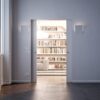 A modern, minimalist room with white walls and large windows opens into a bright library featuring bookshelves, a striped rug, and a light sofa. The ECLISSE Pocket Door System - Syntesis® Double Wiring-Ready enhances the natural light flow throughout.