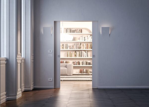 A modern, minimalist room with white walls and large windows opens into a bright library featuring bookshelves, a striped rug, and a light sofa. The ECLISSE Pocket Door System - Syntesis® Double Wiring-Ready enhances the natural light flow throughout.