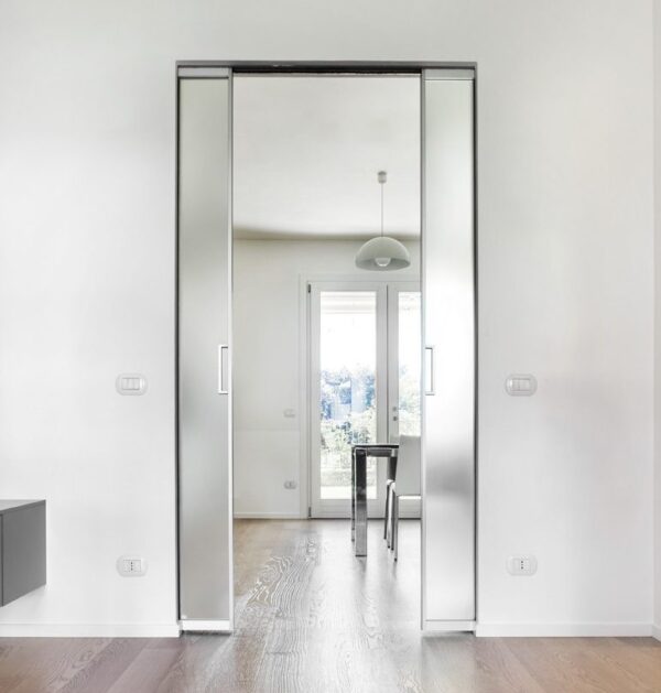 A modern, bright room features the ECLISSE Pocket Door System - Syntesis® Double Wiring-Ready, opening to a dining area with a table, chairs, pendant light, and large windows that fill the space with natural light.