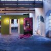 An art gallery in an industrial space features colourful paintings, a green partition with the ECLISSE Pocket Door System—Syntesis® Double Wiring-Ready, two lights, large windows, and pink material scattered on the floor.