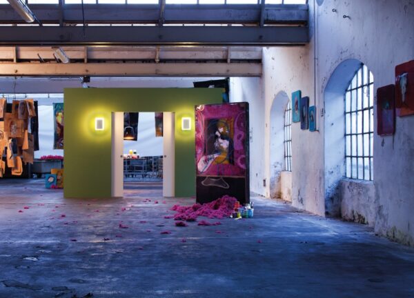An art gallery in an industrial space features colourful paintings, a green partition with the ECLISSE Pocket Door System—Syntesis® Double Wiring-Ready, two lights, large windows, and pink material scattered on the floor.