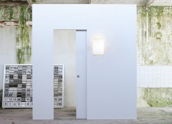 A minimalist white wall with the ECLISSE Pocket Door System - Syntesis® Single Wiring-Ready and a glowing wall light stands in an industrial space, featuring green mould on the back wall and large numbered grids on the left.