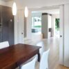 A modern dining space with a wooden table and white chairs sits by a sleek grey kitchen; ECLISSE Pocket Door System - Double Telescopic Sliding opens to a bright living room with large windows and lush greenery views.