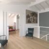 A modern, minimal upstairs landing with light wood floors, white walls, and a white bannister features two dark green chairs by books and a table. A large painting hangs above. The ECLISSE Pocket Door System - Double Telescopic Sliding reveals a bedroom.