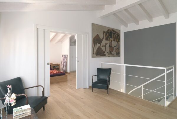 A modern, minimal upstairs landing with light wood floors, white walls, and a white bannister features two dark green chairs by books and a table. A large painting hangs above. The ECLISSE Pocket Door System - Double Telescopic Sliding reveals a bedroom.
